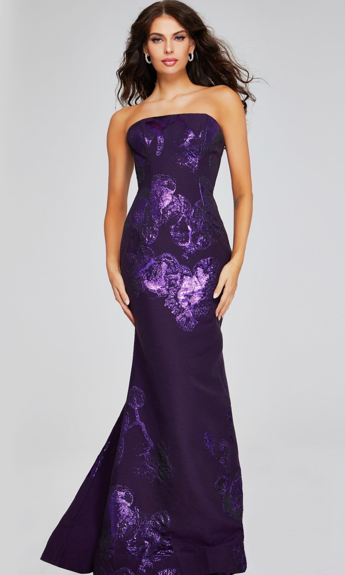 A woman stands against a plain light background wearing the Jovani 40318 long formal dress, a strapless metallic satin mermaid evening gown with a fitted silhouette and subtle floral pattern.